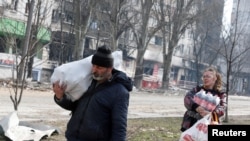 在被俄军围攻的南部港城马里乌波尔，当地居民肩扛手提，携带着食物走过被战火破坏的一座居民楼。(2022年3月31日)